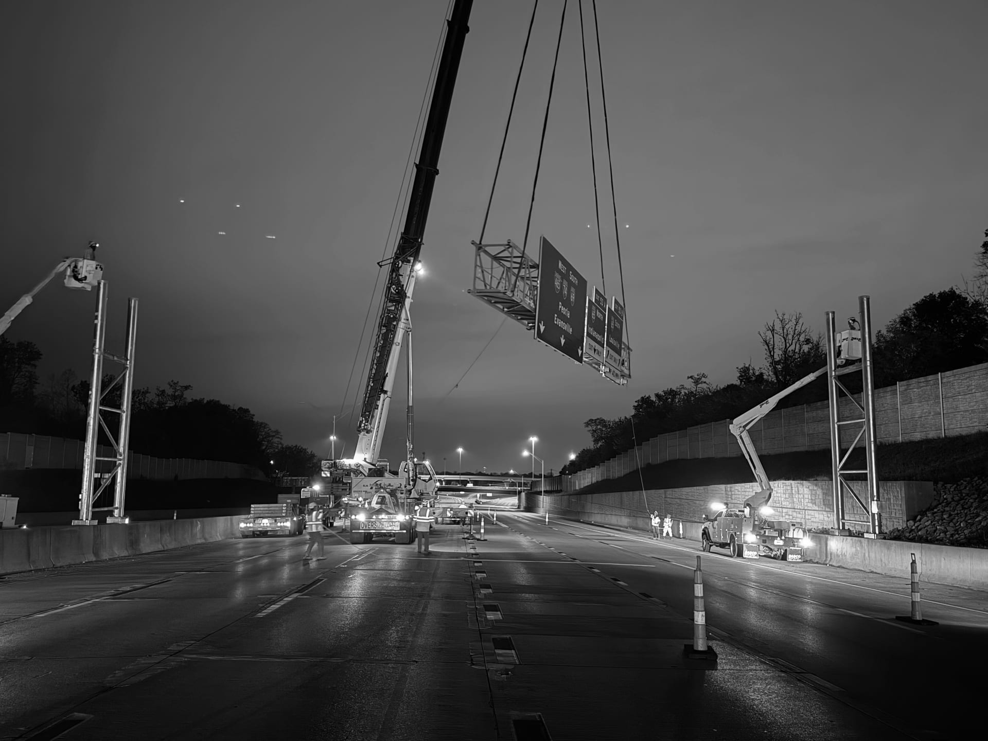 Nighttime installation of an overhead highway sign bridge with a crane lifting a multi-panel sign assembly above an I-465 interchange — James H Drew Corporation sign structure installation